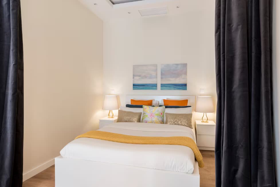 Neatly made double bed with white linen, mustard throw and bedside lamps in a calm short-let bedroom